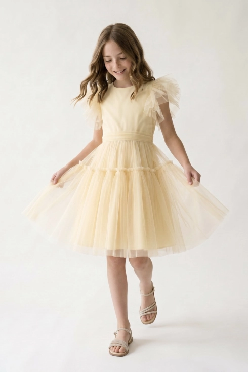 Sukienka tiulowa dla dziewczynki Paola w maślanej żółci — ujęcie z przodu: dziewczynka rozkłada obiema rękami szeroki klosz spódnicy, widoczne piórka przy rękawkach i tiulowy pas w talii